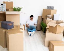 person working on a laptop at home, surrounded by packages and shipping supplies resmi