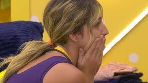 Participante do BBB 23 com a mão no rosto, em momento de reflexão ou preocupação. Ambiente interno com fundo amarelo.