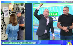 Homem comemora com punho erguido em programa de TV, ao lado de outro homem. Ao fundo, cena de abordagem policial em posto de…