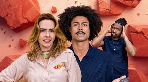 Três pessoas em frente a um fundo com pedras vermelhas quebradas. Uma mulher loira sorri, um homem negro com cabelo afro est…