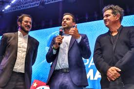 Três homens em um palco, um deles falando ao microfone. Ao fundo, um banner com letras azuis e brancas.