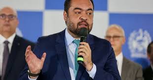 Homem de terno azul e gravata azul discursa em um microfone, com outras pessoas ao fundo.