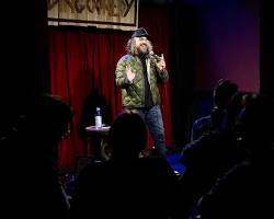 comedian standing in front of a microphone, holding a bag of money.