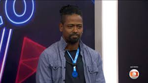 Participante do BBB 23, Fred Nicácio, com barba e cabelo preso, veste camisa azul e medalha no pescoço.