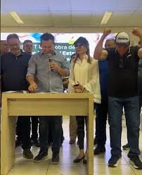 Homem comemorando com braços erguidos ao lado de um homem com microfone e uma mulher com chapéu em evento público.