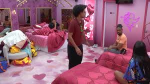 Participantes do BBB 23 em quarto decorado com corações, com camas arrumadas e roupas espalhadas.