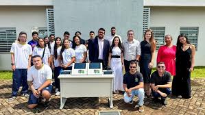 Grupo de pessoas posa para foto em frente a um prédio, com duas urnas eletrônicas em uma mesa à frente.