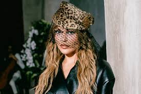 Anitta usa chapéu de oncinha e véu preto em pose misteriosa, com cabelo loiro ondulado e jaqueta de couro.