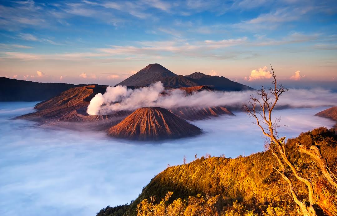 Pemandangan dramatis gunung berapi aktif di Indonesia dengan asap mengepul dari kawahnya, dengan latar belakang lanskap pegunungan yang subur di siang hari.