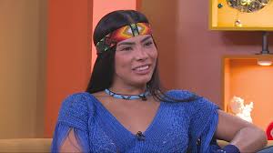 Jaciara Dias sorri usando um colar e uma faixa na cabeça com detalhes coloridos, em um estúdio de TV.