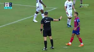 Juiz de futebol impede jogador de avançar em campo durante partida. Placar indica Bahia x Vitória.