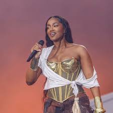 Cantora Tyla em performance com figurino dourado e branco, segurando microfone em palco com fundo alaranjado.