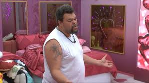 Homem em quarto rosa com cama desarrumada e quadro decorativo na parede. Ele gesticula com as mãos.