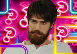 Ator turco Çağatay Ulusoy com barba e bigode, em frente a um fundo rosa com símbolos de interrogação neon.