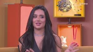 Jovem mulher de cabelos longos e escuros, usando um colar de diamantes, em um estúdio de TV com decoração colorida.