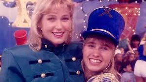 Xuxa Meneghel e Angélica em figurinos de desfile, sorrindo para a câmera em um cenário festivo.