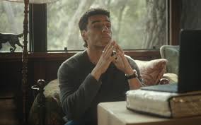 Homem pensativo com as mãos juntas em frente ao rosto, sentado em uma sala com uma janela ao fundo.