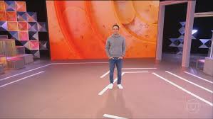 Tadeu Schmidt em um estúdio de TV com um telão ao fundo exibindo uma imagem abstrata em tons de laranja.