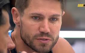 Homem com barba e cabelo curto olha para o lado, com um leve sorriso. Logo da BBB ao fundo.