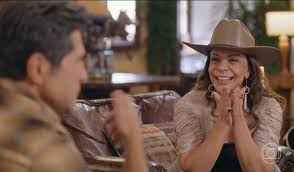 Atriz Maitê Proença sorri usando chapéu country em cena de novela, em conversa com ator não identificado.