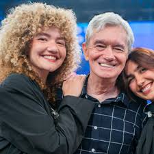 Silvio Santos sorri com suas netas, que o abraçam em um estúdio de TV. As jovens são Rebeca Abravanel e Patrícia Abravanel.